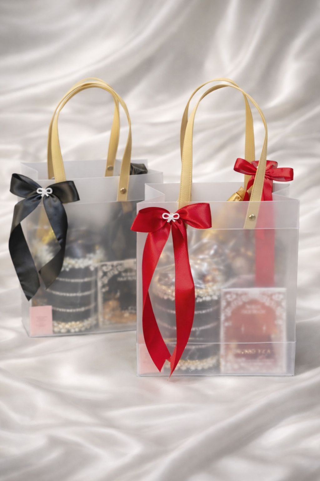 Two clear gift bags with black and red ribbons on a white fabric background