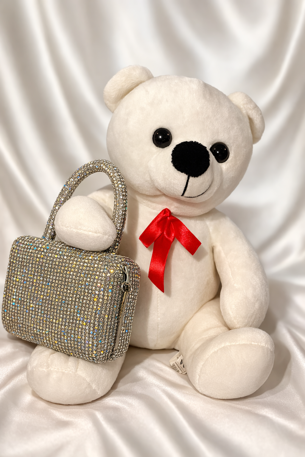 White teddy bear with a red bow and a sparkling handbag on a white satin background